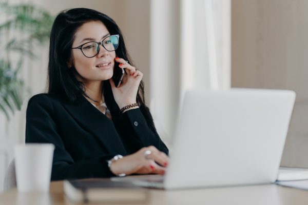 Confident serious business lady has phone conversation, poses in front of laptop computer